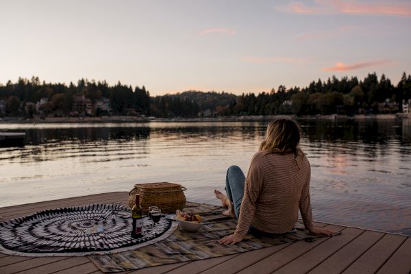 A person sits on a dock by a lake during sunset, with a picnic setup including a basket and snacks on a blanket.