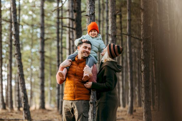 A family enjoying a day in the forest; a man carries a child on his shoulders while a woman stands beside them, with tall trees surrounding them.