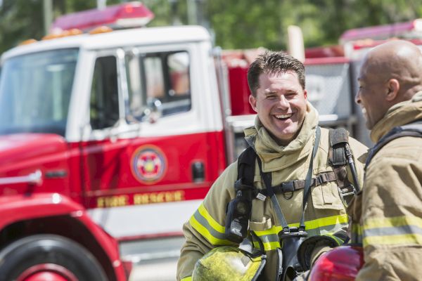 Two firefighters in gear are smiling near a bright red fire truck outdoors, appearing to have a casual conversation.