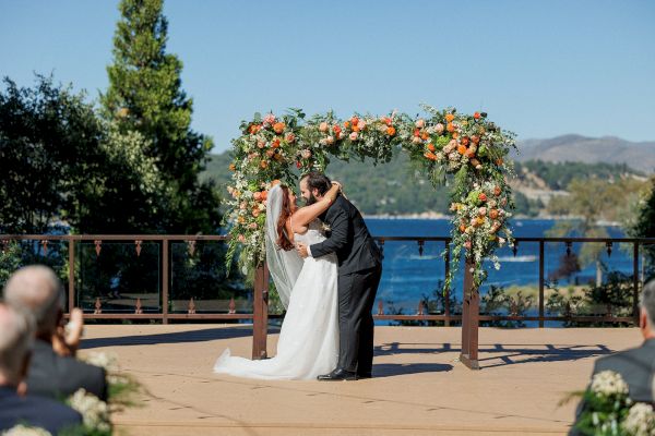 A couple is kissing under a floral arch at an outdoor wedding ceremony with a scenic background of trees and a body of water.