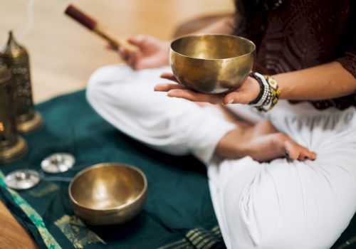 A person sits cross-legged on a mat, holding a singing bowl with another bowl nearby and lanterns in the background, creating a peaceful atmosphere.