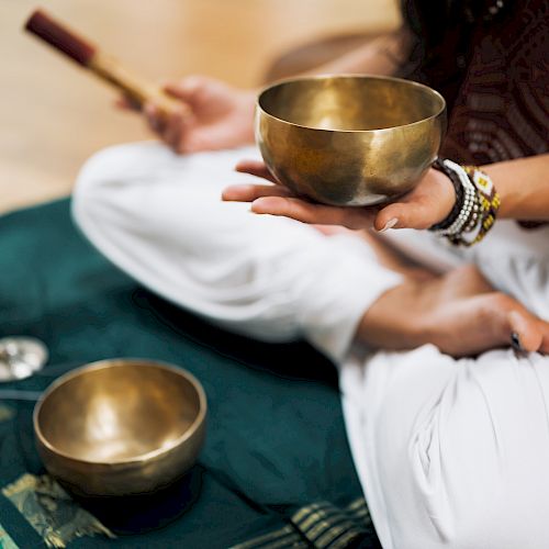 A person sits cross-legged on a mat, holding a singing bowl with another bowl nearby and lanterns in the background, creating a peaceful atmosphere.