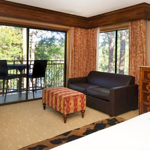 A cozy room with a sofa, ottoman, TV, and a balcony overlooking trees.