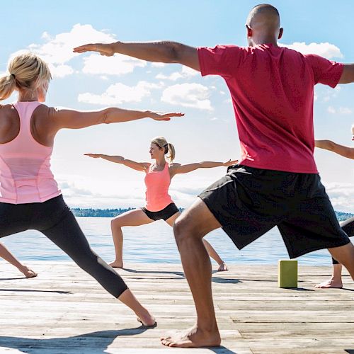 Group of people practicing yoga on a dock by a lake, doing a warrior pose.