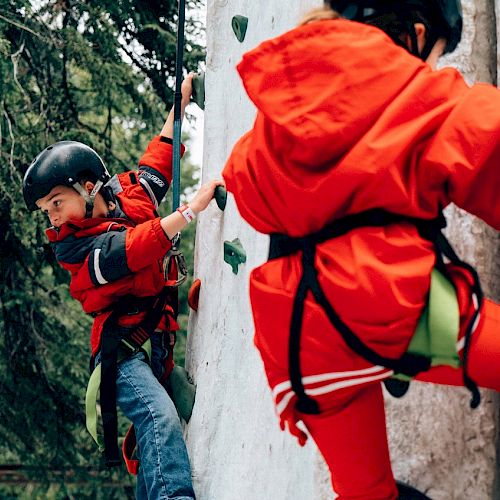 Two children in red climbing gear are ascending an artificial climbing wall outdoors.