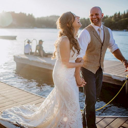 A couple in wedding attire is laughing on a dock at sunset, with boats and water in the background.