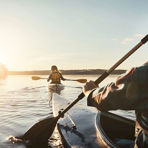 Two people are kayaking on calm waters with the sun setting in the distance.