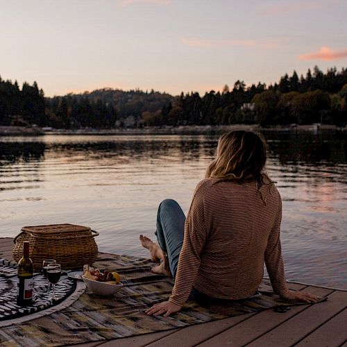 A person sits on a dock by Lake Arrowhead at dusk with a picnic setup, gazing at the serene water.