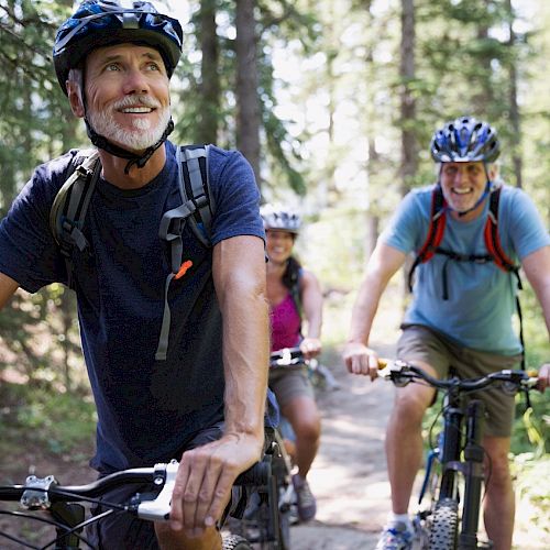 Three people are mountain biking through a forest, wearing helmets and backpacks, looking happy.