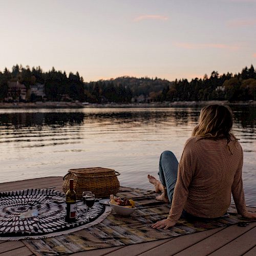 A person sits by a water body at dusk with a picnic setup on a dock.