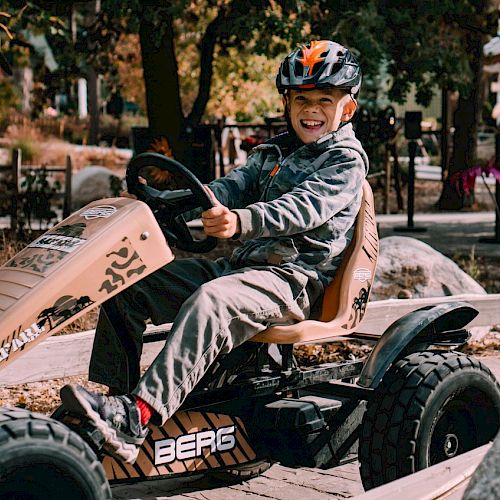 A child is driving a pedal go-kart outdoors, wearing a helmet and smiling.