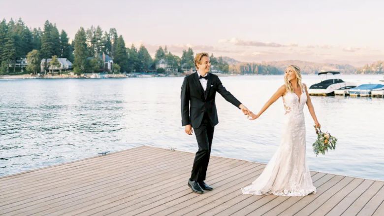 A couple in wedding attire holds hands on a dock by the water at sunset.