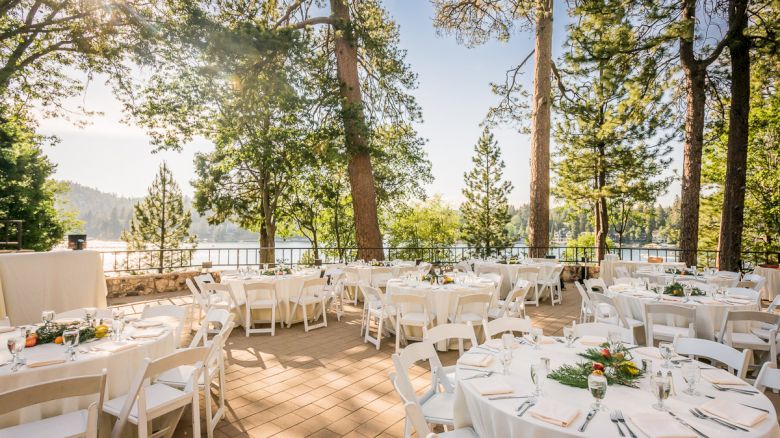 Outdoor event setting with white chairs and tables, near trees with a lake view.