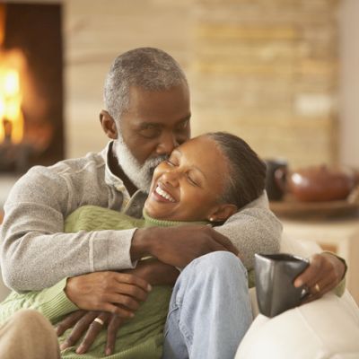 A couple is hugging on a couch with a fireplace in the background.