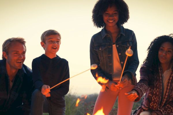 A group is toasting marshmallows over a fire at dusk, sharing a joyful moment.