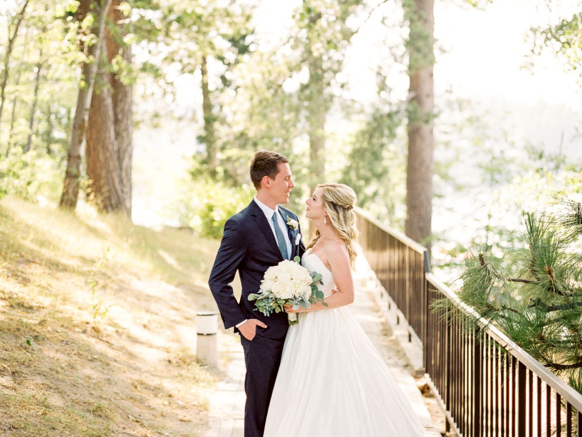 A couple in wedding attire is standing by a fence, surrounded by trees in sunlight.