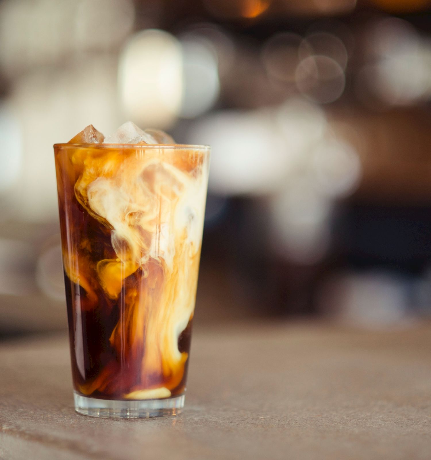 Iced coffee with cream swirling beautifully in a glass, on a blurry background.