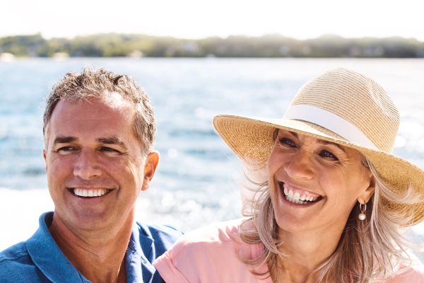 Two people are smiling on a boat, with water and sunlight in the background.