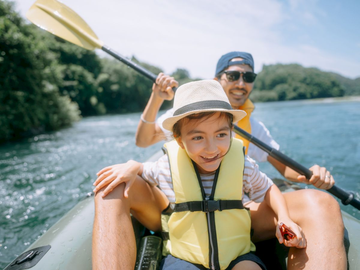 Two people are kayaking on a river, one in front is smiling, and both wear life jackets.