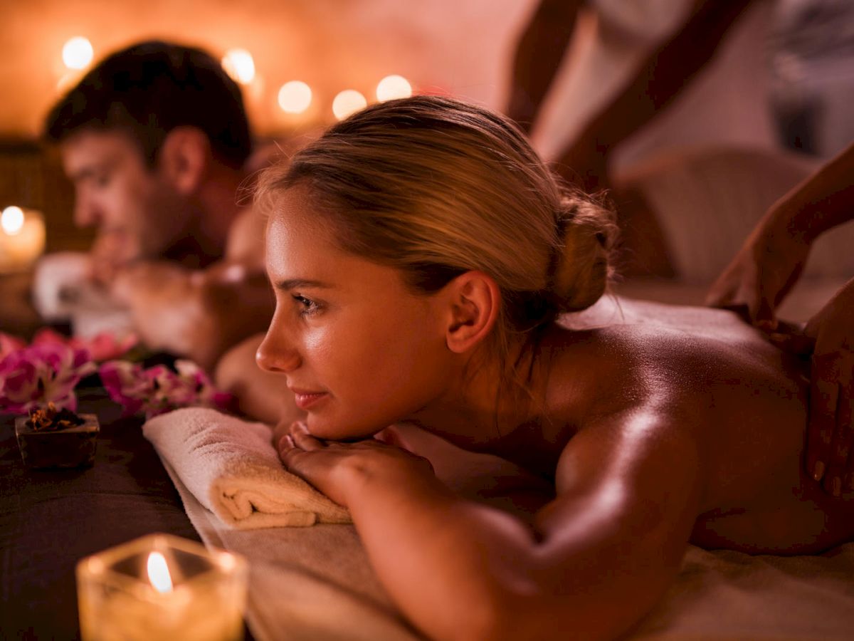 Two people getting a couples massage in a dimly lit spa treatment room surrounded by lit candles.