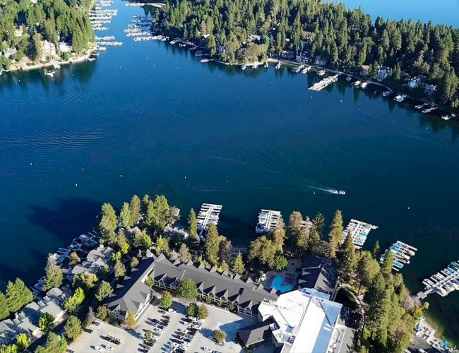 Aerial view of a serene lake surrounded by dense forests, with numerous docks and boats, and several buildings located near the water's edge.