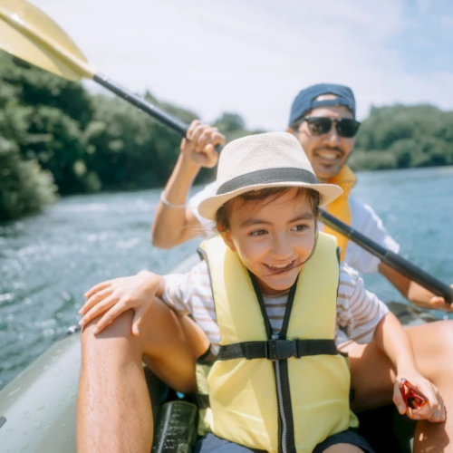 A child and an adult are smiling while kayaking on a river, both wearing life vests, on a sunny day, surrounded by lush greenery.