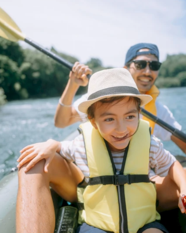 A child and an adult are smiling while kayaking on a river, both wearing life vests, on a sunny day, surrounded by lush greenery.