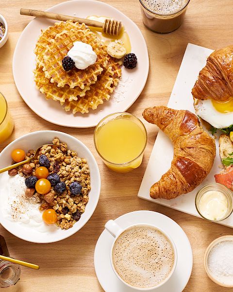 A variety of breakfast items are on the table, including waffles, croissants, eggs, yogurt with granola, pastries, juices, and coffee.