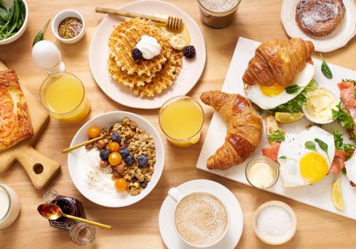 A variety of breakfast items are on the table, including waffles, croissants, eggs, yogurt with granola, pastries, juices, and coffee.