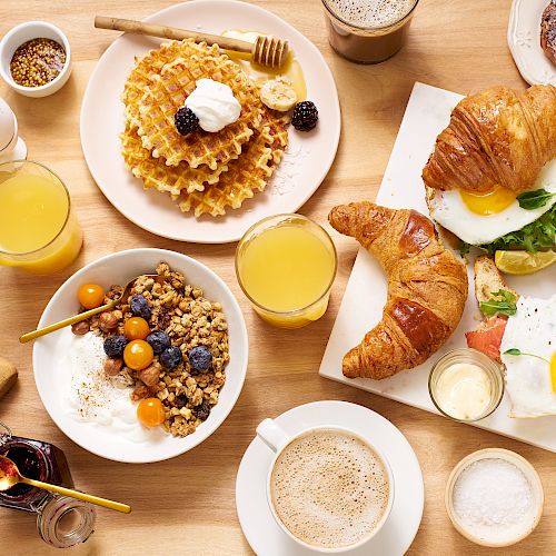 A variety of breakfast items are on the table, including waffles, croissants, eggs, yogurt with granola, pastries, juices, and coffee.