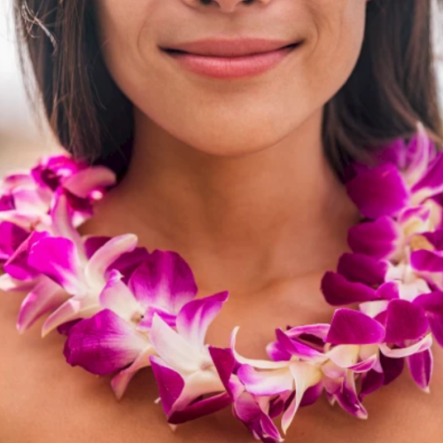 A close-up image of a person smiling, wearing a vibrant purple and white flower lei around their neck.