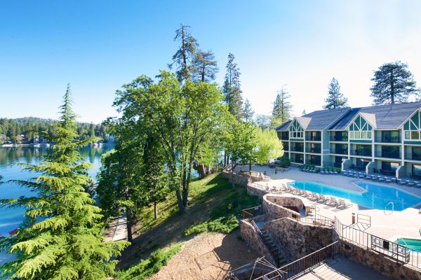 A lakeside resort with a pool, surrounded by trees, features modern buildings under clear blue skies and looks serene and inviting.