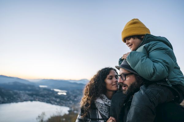 A family enjoys a scenic view from a hilltop at dusk, with a child on one adult's shoulders and the others standing beside, overlooking a city.