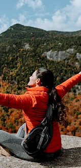 A person in an orange jacket sits on a rock ledge, arms outstretched, overlooking a scenic view of trees and a river with mountains.