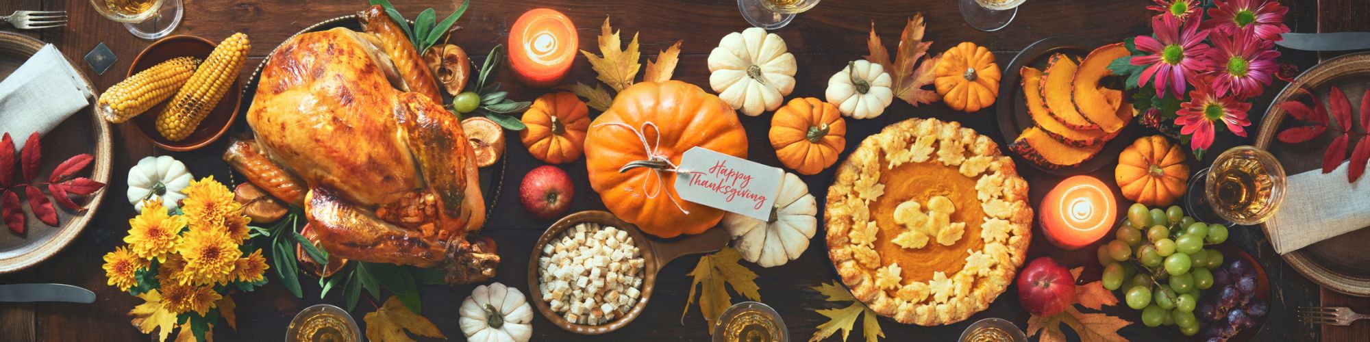 A festive table set for a Thanksgiving meal with a roasted turkey, pumpkin pie, fall leaves, small pumpkins, corn, grapes, and candles.