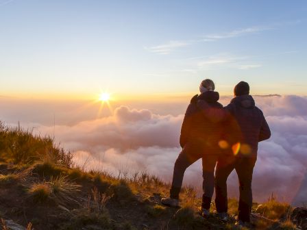 Two people stand on a mountaintop, watching a sunrise above the clouds, surrounded by grass and enjoying the scenic view.