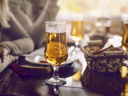 A glass of beer on a table with partially visible people and a breadbasket nearby, creating a casual dining atmosphere.