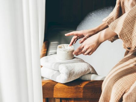 A person in a robe applying cream from a jar beside folded towels near a bubble-filled bathtub, creating a calming spa-like atmosphere.