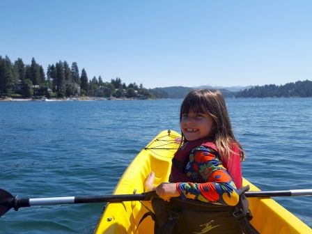 A child smiles while kayaking on a calm lake with a forested shoreline in the background under a clear blue sky.