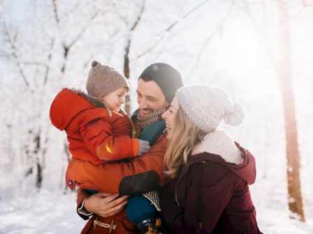 A family of three in winter clothing is enjoying a snowy day outdoors, with snow-covered trees in the background.