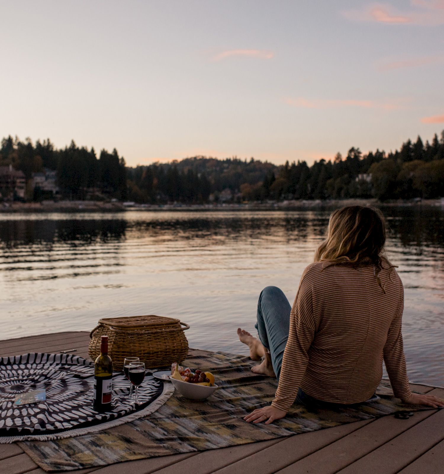 A person sits on a dock by a lake at sunset, next to a blanket, a basket, and snacks, enjoying the serene view of the water and trees.