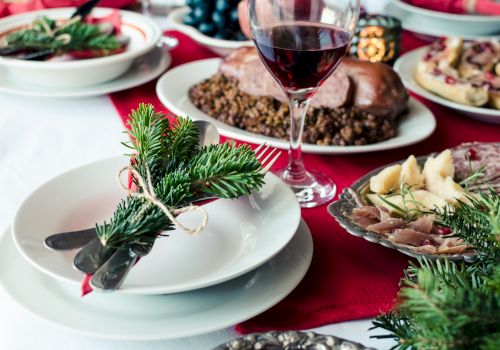 A festive table setting with a plate, cutlery wrapped in greenery, various dishes, and a glass of red wine on a red cloth.