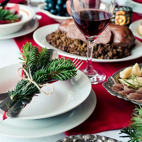 A festive table setting with a plate, cutlery wrapped in greenery, various dishes, and a glass of red wine on a red cloth.