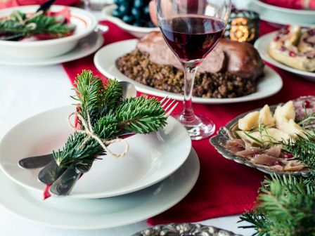 A festive table setting with pine decoration, dishes with food, a glass of red wine, and a red tablecloth.