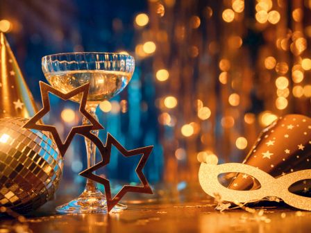 A festive celebration scene with a champagne glass, star-shaped glasses, party hats, a disco ball, and a masquerade mask, with lights.