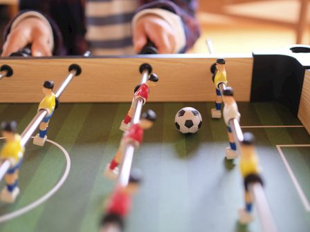 The image shows a foosball table with players in yellow and red uniforms, a ball in the center, and someone playing.