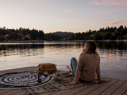 A person sits on a lakeside dock with a picnic setup, enjoying a tranquil sunset view over the water, surrounded by trees.