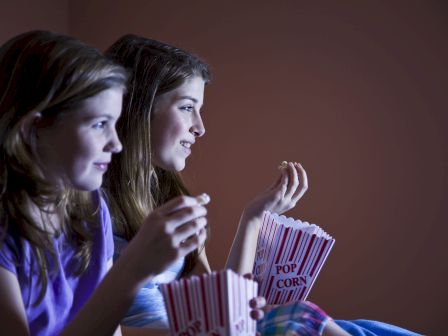 Two people are watching something, holding popcorn boxes, in a dimly lit room.