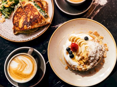 A table with a cappuccino, toasted sandwich with salad, and a dessert topped with berries, almonds, and cream.