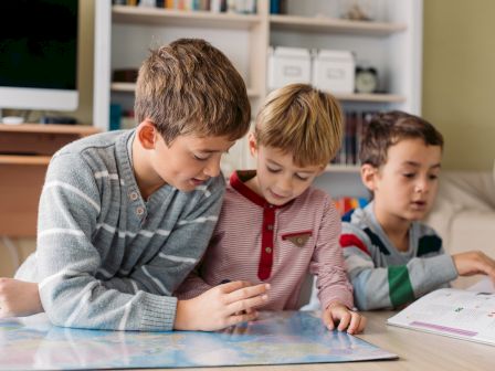 Three children are looking at a map together in a room, with books and a computer in the background.